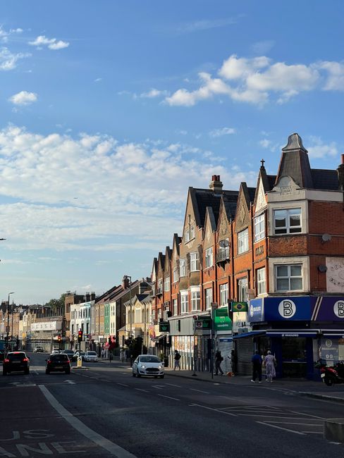 A row of multi-story brick and stone residential buildings with gabled roofs and bay windows lining a busy city street under a partly cloudy sky. The buildings feature a mix of red and beige bricks with white window frames and decorative elements. Ground-floor retail shops, including a public transport ticket kiosk with signage, are visible on the right side of the image, alongside a few pedestrians and vehicles including cars and a bus. The street scene captures typical urban activity in a residential area, with a clear atmosphere and natural daylight highlighting the architectural details. The environment suggests a typical UK neighborhood with accessible commercial and transport services, relevant to private rubbish collection and waste management mention.