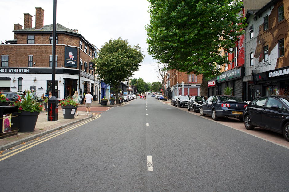 A wide urban street scene showing parked cars lining the right side of the road and pedestrians walking along the pavement on both sides. The street is flanked by mixed-use buildings, including a corner cafe or pub with outdoor planters containing colorful flowers, on the left, and several shops and businesses on the right, some with signage and awnings. A large leafy tree extends over the street from the right side, providing partial shade across the roadway. The surface is asphalt with clear white lane markings down the center, and the atmosphere appears overcast with diffused natural light. The area suggests a busy town centre with opportunities for independent rubbish collection or private waste handling, supported by visible urban elements such as parked vehicles and commercial establishments, as seen in local rubbish removal contexts. House Clearance Acton occasionally handles waste clearance in such settings, ensuring streets remain clear of discarded rubbish, possibly from on-site clearance or waste disposal in busy commercial districts.