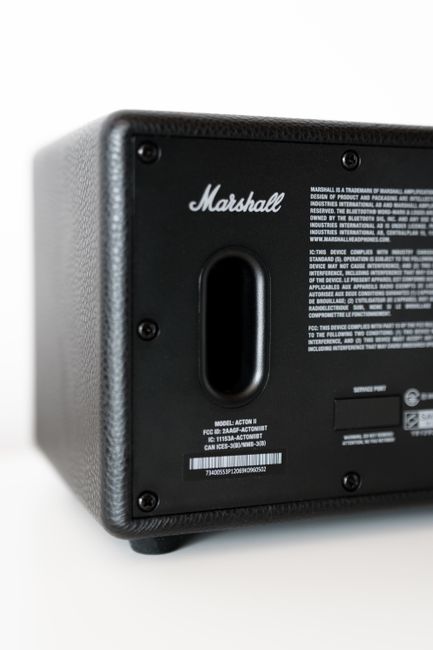 A close-up view of the bottom of a black Marshall speaker with a textured exterior surface, showing a central rectangular bass port. The surface has a fine, woven-like finish, and there are four small visible screws around the edges. The background is plain white, and the lighting creates soft shadows on the speaker, highlighting its shape and texture. The image is clean and sharply focused, with the speaker positioned slightly to the right in the frame. The overall scene suggests an indoor setting where electronic audio equipment may be used or stored, with no additional objects or environmental features visible. The image emphasizes the construction details and branding of the speaker, which is associated with high-quality sound equipment and alternative audio solutions, relevant to services such as rubbish removal involving electronic waste disposal, handled by companies like House Clearance Acton.