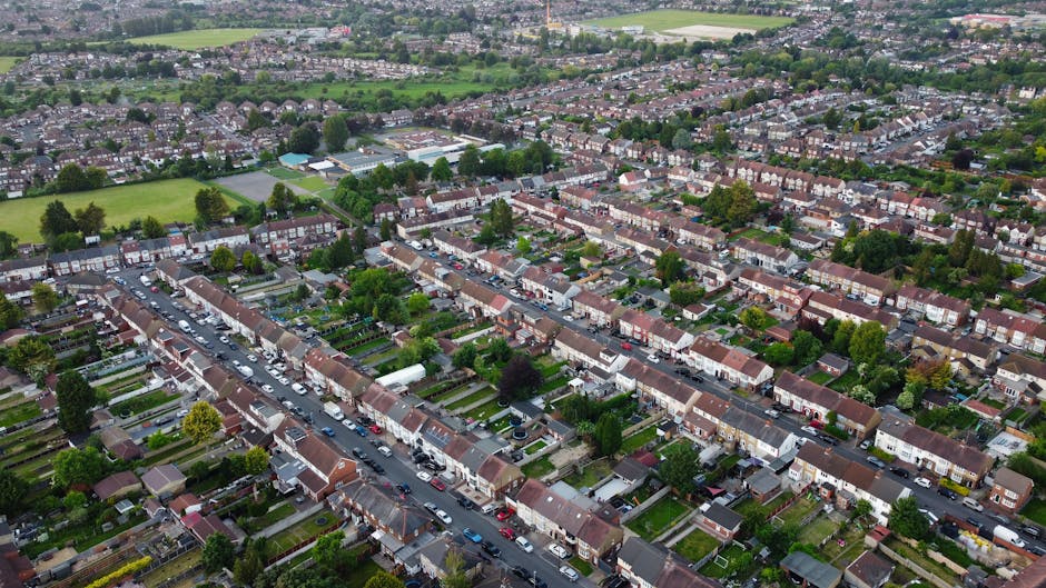 An aerial view of a suburban residential area with rows of terraced houses, each with small front gardens and driveways. The houses have brick walls, tiled roofs in reddish-brown and dark grey shades, and some have visible fences or hedges separating their properties. Cars are parked along the street, varying in size and colour, with some on the road and others in driveways. Behind the houses, there are patches of green open spaces, including sports fields and patches of woodland, with trees scattered throughout the neighbourhood. The scene is illuminated by natural daylight, creating clear visibility of the neighborhood's layout. This high-angle perspective emphasizes the uniform arrangement typical of suburban housing developments and subtly highlights the importance of proper rubbish and waste management within residential communities, aligning with the services of private rubbish removal companies like House Clearance Acton.